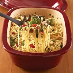 a red casserole dish with broccoli and noodles in it on a wooden table