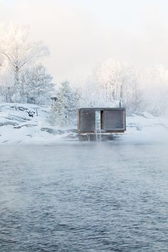 a body of water surrounded by trees and snow covered ground with two buildings on the side