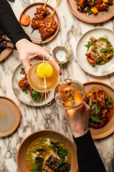 two people are toasting with drinks at a table full of plates and bowls filled with food