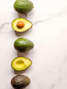 avocados are arranged in a row on a white marble countertop, with one cut in half and the other whole