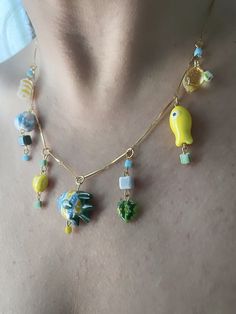 Capture the essence of a European summer with this handmade charm necklace, adorned with ceramic and glass beads on a gold-plated chain. Inspired by seaside getaways, it showcases ceramic fish charms, millefiori-like glass beads, a glass lemon charm, and a diverse array of other ceramic and glass beads. This color way is green, yellow and aqua blue. The bar chain, links, and clasp are all elegantly plated in 18k or 24k gold. For longevity, refrain from exposing this jewelry to water, lotions, perfume, or similar substances to prevent tarnishing. Ceramic Fish, Handmade Charms, European Summer, Glass Necklace, Aqua Blue, Charm Necklace, Glass Beads, Beaded Necklace, Jewelry Necklaces