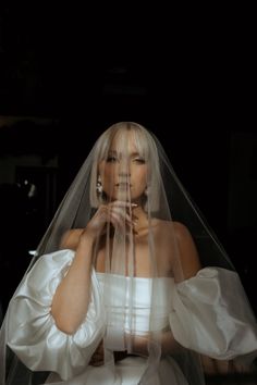 a woman wearing a white wedding dress and veil with her hands on her face, standing in front of a black background