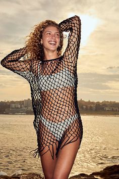 a woman in a fishnet swimsuit standing on the beach with her arms behind her head