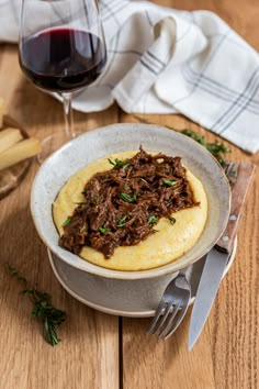 a bowl filled with food next to a fork and glass of wine on top of a wooden table