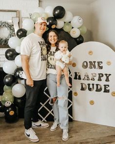 a man and woman holding a baby in front of a balloon arch with the words one happy dude on it