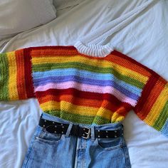 a rainbow sweater and jean shorts on a bed with white sheets in the background as well as a pair of black belted jeans