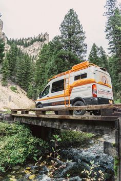 an orange and white van driving over a bridge in the woods next to some trees