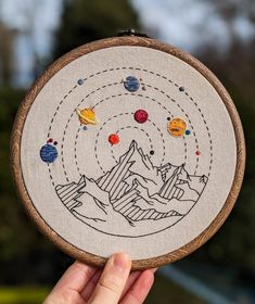 a hand holding up a small embroidery project with planets and mountains in the sky on it