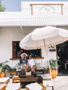 two people sitting under an umbrella in front of a building with potted plants on the outside