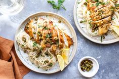 two plates of pasta with chicken, capers and lemon wedges on the side