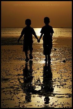 two children holding hands while walking on the beach at sunset or dawn with water reflecting off the sand