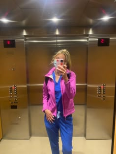 a woman taking a selfie in front of an elevator with blue pants and pink jacket