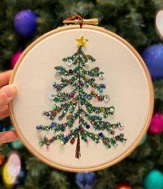 someone holding up a cross stitch christmas tree ornament in front of a decorated christmas tree