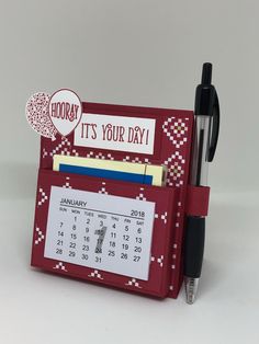 a red desk calendar holder with a pen and pencil in it that says, it's your day