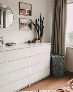 a white dresser sitting in front of a window next to a mirror and potted plant