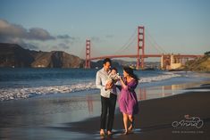 a man and woman are walking on the beach with a baby in their arms near the golden gate bridge