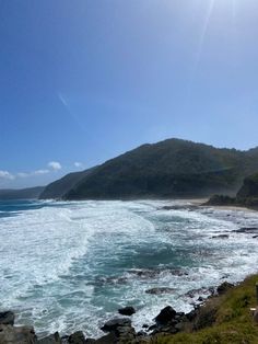 an ocean view with waves crashing on the shore and mountains in the distance, under a bright blue sky