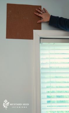 a person holding up a piece of cardboard on top of a window sill next to blinds