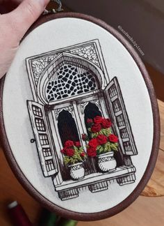 a person holding up a small embroidery project with red flowers in front of an open window