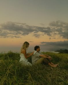 a man and woman sitting on top of a lush green field next to the ocean