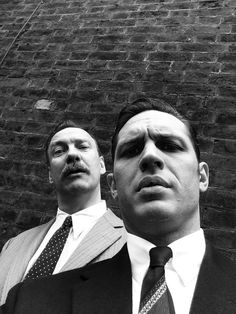 two men standing next to each other in front of a brick wall wearing suits and ties