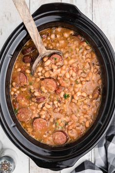 a black crock pot filled with beans and sausages next to a wooden spoon