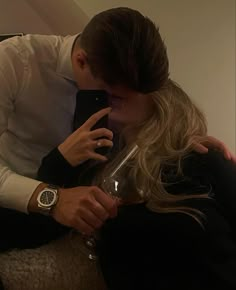 a man and woman sitting on a couch kissing while looking at their cell phones with wine glasses in front of them