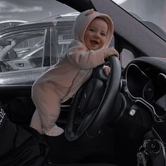 a baby is sitting in the driver's seat of a car, wearing a hoodie