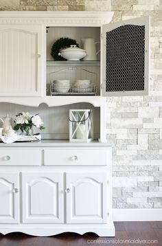 a white china cabinet with flowers and plates on it's shelves in front of a brick wall