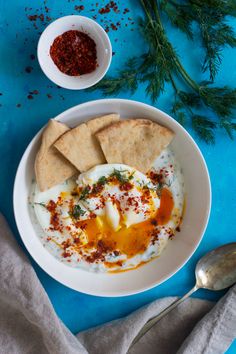 an egg and pita on a white plate next to a bowl of sauce, spoons and napkin