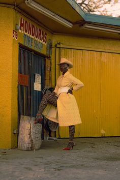 a woman in a yellow coat and hat leaning against a door with her legs crossed