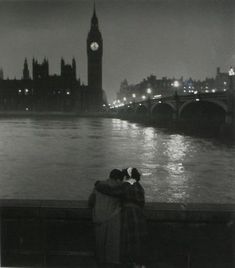 two people standing next to each other on a bridge near the water with a clock tower in the background