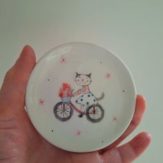 a hand holding a white plate with a cat riding a bike on it's side