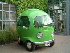 a small green van parked in front of a building with plants growing on it's side