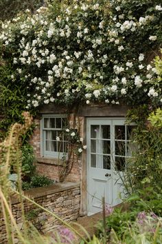 a house with white flowers growing on it's roof and door, surrounded by greenery