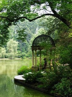 a gazebo in the middle of a lake surrounded by trees and bushes with a man sitting on it