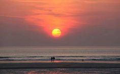 two people are walking on the beach as the sun sets in the distance behind them