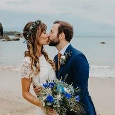 a bride and groom kissing on the beach with blue flowers in their hair, dressed in a navy suit