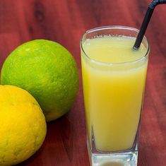 a glass of orange juice next to two limes on a wooden table with a black straw