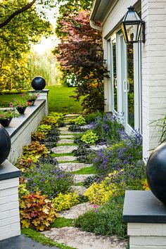 an outdoor garden with flowers and plants on the side of a white brick building next to a walkway