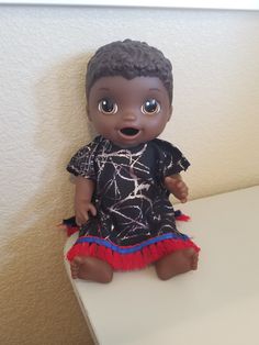 a small doll sitting on top of a white table next to a wall with a black shirt and red skirt