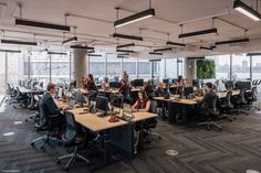 an office filled with people sitting at desks and working on their computers in front of large windows