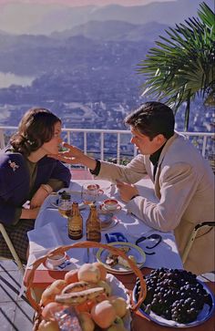 a man and woman sitting at a table with food in front of them on a balcony