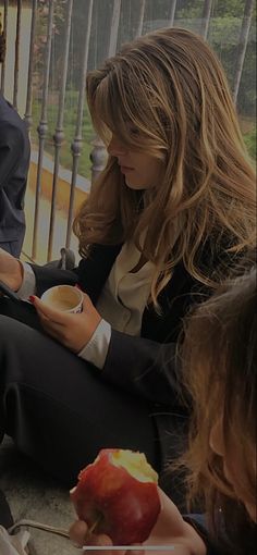 a woman sitting on the ground holding an apple and looking at her cell phone with another person standing next to her