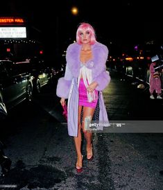 a woman with pink hair is walking down the street at night wearing a purple fur coat