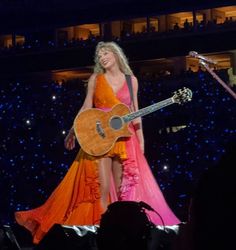 a woman in an orange and pink dress holding a guitar while standing on top of a stage