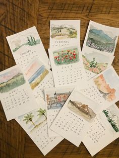 several calendars with watercolor paintings on them sitting on top of a wooden table