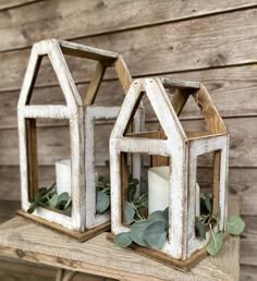 two white wooden lanterns with greenery and candles on a wood shelf next to a wall