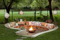 an outdoor fire pit surrounded by trees and grass with lights hanging from the branches above it