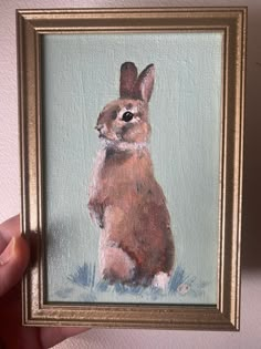 a painting of a brown rabbit in a gold frame on a white wall with a hand holding it up